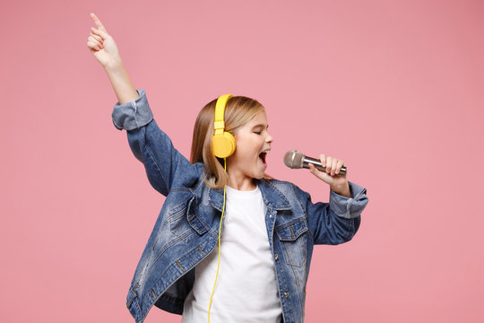 Cute little kid girl 12-13 years old isolated on pastel pink background. Childhood lifestyle concept. Mock up copy space. Listen music with headphones sing song in microphone pointing index finger up.