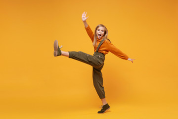 Fototapeta na wymiar Cheerful little blonde kid girl 12-13 years old in turtleneck, jumpsuit isolated on orange yellow background. Childhood lifestyle concept. Mock up copy space. Dancing, rising leg, spreading hands.