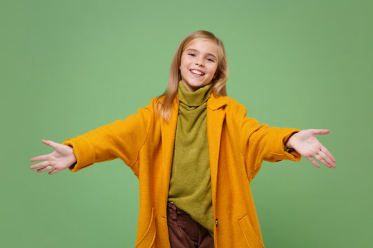 Pleasant little blonde kid girl 12-13 years old in yellow coat posing isolated on pastel green background in studio. Childhood lifestyle concept. Mock up copy space. Standing with outstretched hands.