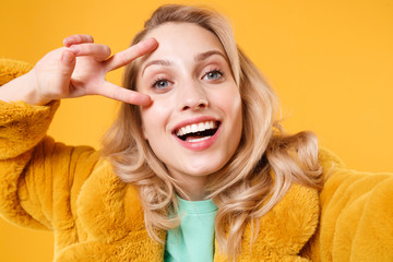 Close up of excited young blonde woman in yellow fur coat posing isolated on orange background. People lifestyle concept. Mock up copy space. Doing selfie shot on mobile phone, showing victory sign.