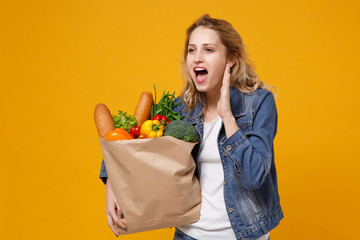 Woman in denim clothes isolated on orange background. Delivery service from shop or restaurant concept. Hold brown craft paper bag for takeaway mock up with food products screaming with hand gesture.