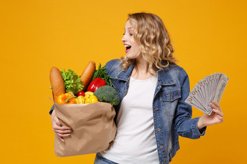 Amazed young girl in denim clothes isolated on orange background. Delivery service from shop or restaurant concept. Hold brown craft paper bag for takeaway mock up with food product fan of cash money.