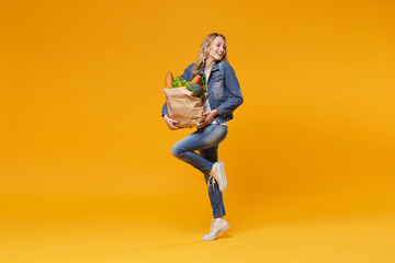 Fototapeta na wymiar Cheerful young woman in denim clothes isolated on yellow orange wall background. Delivery service from shop or restaurant concept. Hold brown craft paper bag for takeaway mock up with food products.