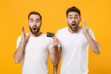 Shocked young men guys friends in white blank empty t-shirts posing isolated on yellow orange background in studio. People lifestyle concept. Mock up copy space. Hold credit bank card spreading hands.