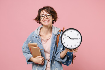 Cheerful young woman student in denim clothes eyeglasses, backpack posing isolated on pastel pink background. Education in high school university college concept. Mock up copy space. Hold books clock.