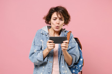 Worried young girl student in denim clothes backpack isolated on pastel pink background in studio. Education in high school university college concept. Mock up copy space. Play game with mobile phone.