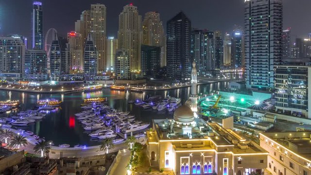 Yachts in Dubai Marina flanked by the Al Rahim Mosque and residential towers and skyscrapers aerial timelapse during all night with lights switching off. Modern high rise skyline with boats and wooden