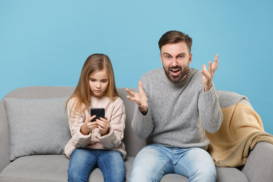 Angry Bearded Man In Knitted Sweater With Child Baby Girl. Father Little Daughter Isolated On Pastel Blue Background. Love Family Parenthood Childhood Concept. Sitting On Couch, Hold Mobile Phone.