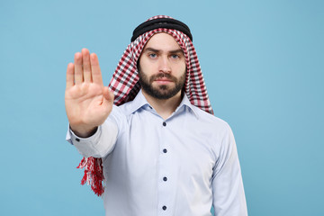 Serious young bearded arabian muslim man in keffiyeh kafiya ring igal agal casual clothes posing isolated on pastel blue background. People religious lifestyle concept. Showing stop gesture with palm.