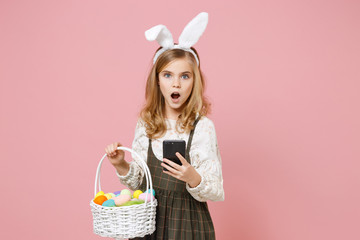 Little blonde kid girl 11-12 years old in spring dress bunny rabbit ears hold in hand cell phone wicker basket colorful eggs isolated on pastel pink background Childhood lifestyle Happy Easter concept