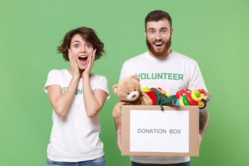 Surprised friends couple in volunteer t-shirt isolated on green background. Voluntary free work assistance help charity grace teamwork concept. Hold donation box with kids toys, put hands on cheeks.