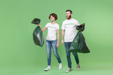 Cheerful two young friends couple in white volunteer t-shirt isolated on pastel green background. Voluntary free work assistance help charity grace teamwork concept. Holding trash bags, looking aside.