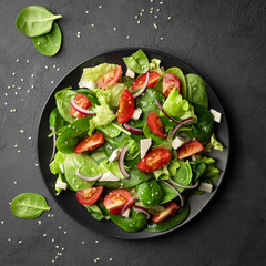 Healthy food. Top view of a fresh green vegetable salad of spinach, tomato, lettuce and sesame seeds on a plate. Black background.