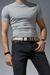 Cropped shot of a man in black jeans and a gray t-shirt on the gray background. A beige canvas belt...