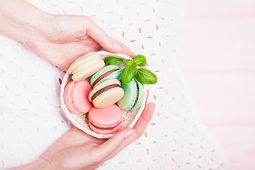 Beautiful female hands holding bowl of french colorful macaroons on white and pale pink background. Top view, copy space