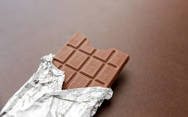 sweets, confectionery and food concept - milk chocolate bar in foil wrapper on brown background