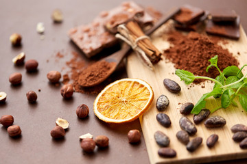 confectionery and culinary concept - chocolate with hazelnuts, cocoa beans, and powder, dry orange and cinnamon on wooden board with spoon on brown background