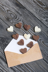 An open white paper envelope on brushed pine boards. Chocolate hearts are scattered from it. From white and black chocolate.