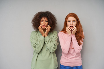 Studio photo of pretty puzzled young ladies holding their faces with raised hands while looking confusedly at camera, isolated over grey background in casual wear
