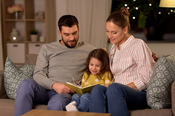 family, leisure and people concept - happy father, mother and little daughter reading book at home at night