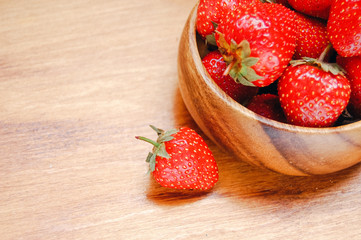 Macro photo of sweet red strawberries in wooden bowl with one berry out at light wooden background.