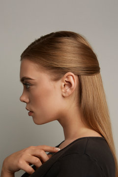 A blonde European girl with long straight hair in a half-up and half-down hairdo is posing on the gray background. The lady is wearing a black tee-shirt. 