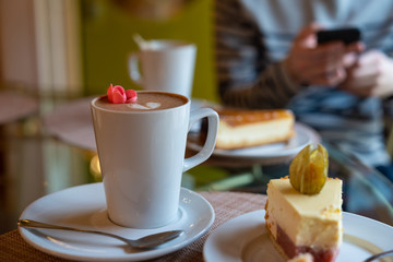Close up of white cup of coffee latte decorated with a heart and flower, cake with bite taken. Man holding smart phone on background. Stock photo.