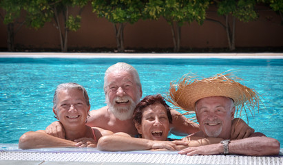 Four happy friends, senior men and women, enjoying the sun in the swimming pool. They laugh looking at the camera
