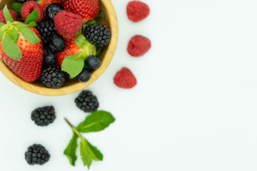 Top view of a wooden bowl filled with strawberries, blackberries, raspberries and blueberries in the upper left corner with fruit on the outside. White background, space for text. Healthy food.