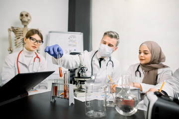 Medicine, genetics, immunology, microbiology concept. Multiethnic group of scientists, Caucasian man and woman, Muslim woman, working at the laboratory with microscope, tubes with liquids and laptop.