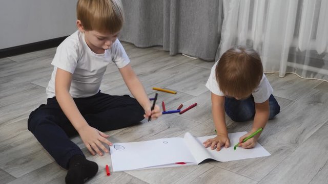 Small blond children sit on the floor and draw on white paper, color with colored pencils.