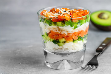 Delicious layered rice salad with salted salmon (trout), cucumber, avocado, cream cheese, black and white sesame seeds in a glass on a gray concrete background. Sushi. Selective focus, copy space