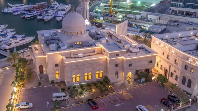 Yachts in Dubai Marina flanked by the Al Rahim Mosque with car parking and residential towers aerial night to day transition timelapse. Modern high rise skyline with boats and wooden dhows