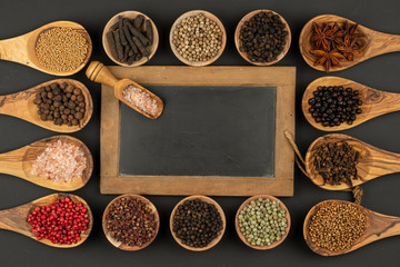 Six small wooden bowls, eight cooking spoons made of olive wood filled with various spices and an old black slate board with copy space lie on a black background