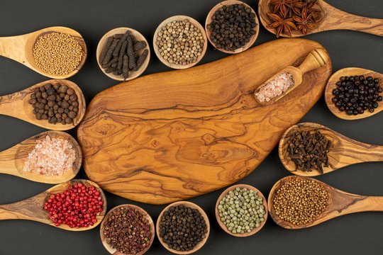 Six small wooden bowls, eight cooking spoons made of olive wood filled with various spices and a wooden spice shovel with pink crystal salt with copy space lie on a black background
