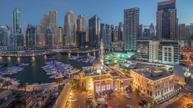 Yachts in Dubai Marina flanked by the Al Rahim Mosque and residential towers and skyscrapers aerial night to day transition timelapse. Modern high rise skyline with boats and wooden dhows