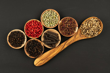 A cooking spoon made of olive wood and six small wooden bowls filled with various peppercorns on a black background - Powered by Adobe