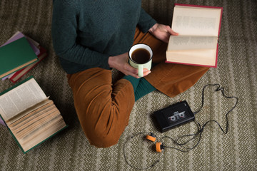 Young student is sitting on a carpet and reading a book, holding a cup of tea. Cassette player for listening a music © Sensay
