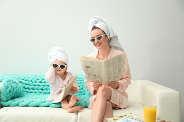Mother and little daughter in bathrobes with newspapers sitting on sofa