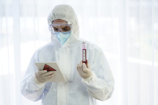 Doctor in personal protective suit looking at a tablet computer and holding the Coronavirus COVID 19 blood sample tube