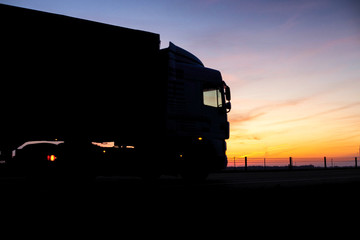 A truck with a trailer rides on the motorway at night against the backdrop of an orange sunny...