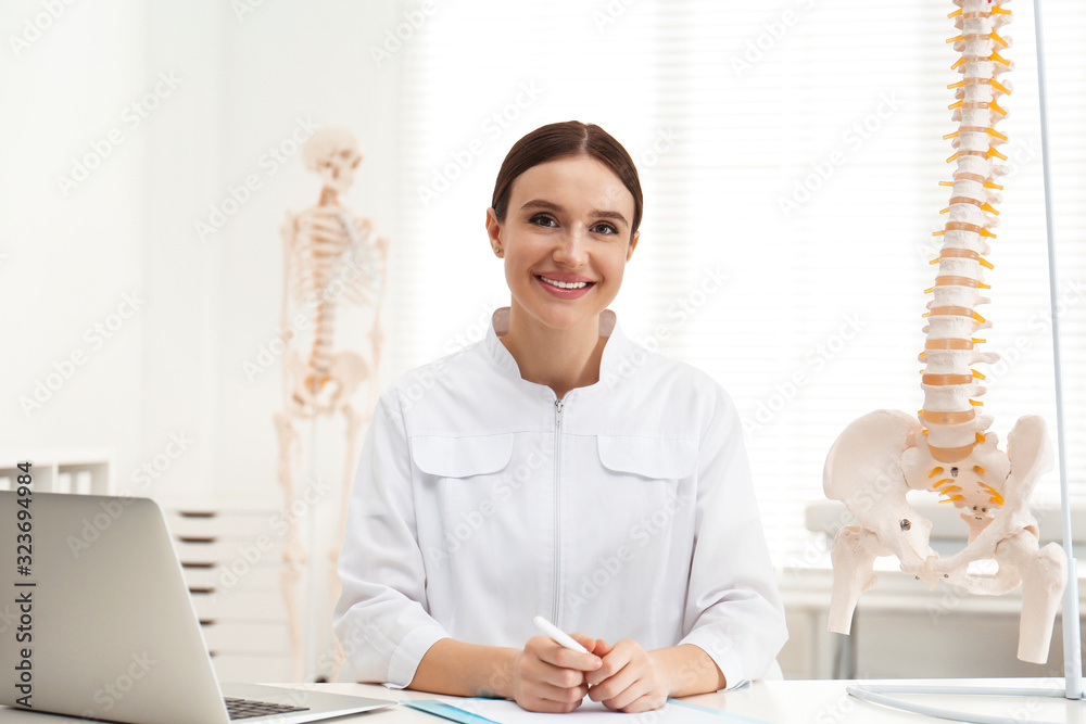 Poster Female orthopedist with laptop near human spine model in office