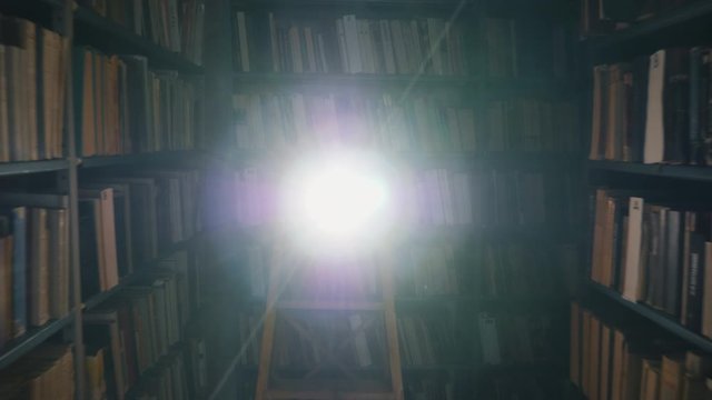 Back move of the rolling camera between rows of bookshelves in a library. The camera moves out of a beam of bright light on the center scene. Indoor