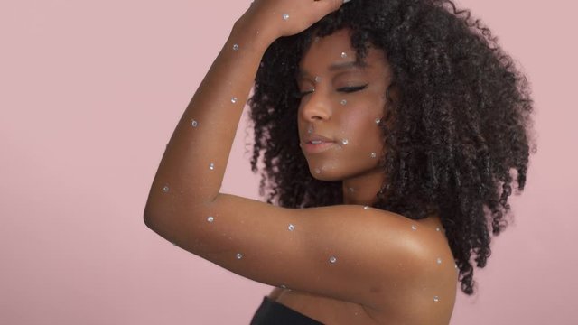 Mixed race black woman with curly hair covered by crystal makeup on pink background in studio Model moves her shoulder dancing