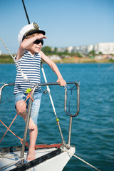 active little caucasian boy in a captain hat and sunglasses peers into distance standing on board of yacht during the summer sea cruise