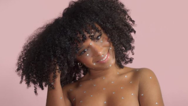 Mixed race black woman with curly hair covered by crystal makeup on pink background in studio Close up smiling and touching her hair dancing
