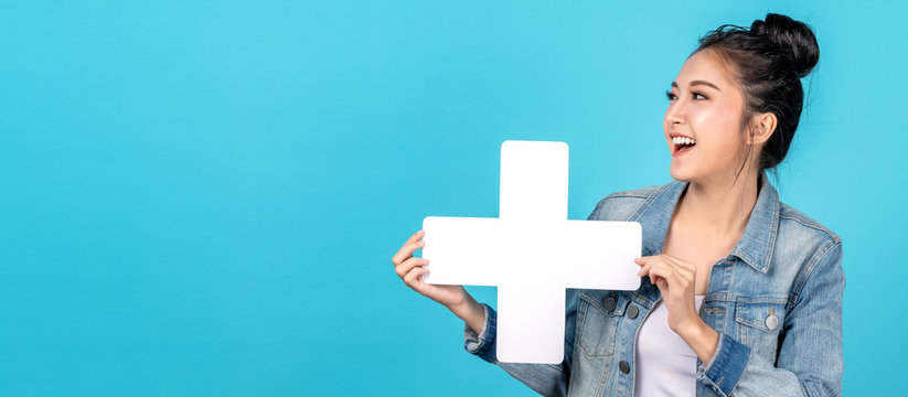 Banner of Happy asian woman smiling and holding plus or add sign on copy space blue background. Cute asia girl smiling wearing casual jeans shirt and showing join sign for increse and more concept