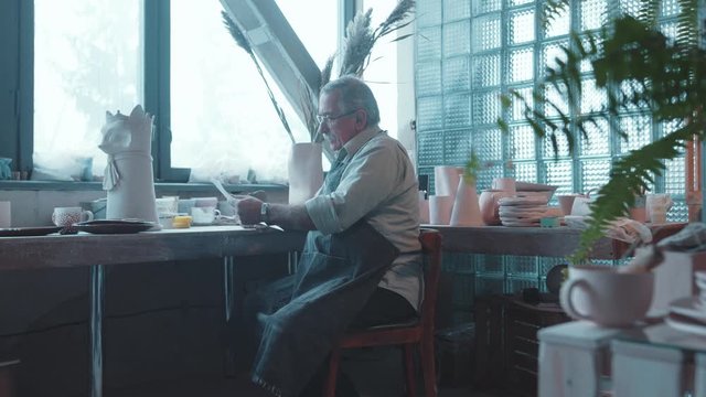 Happy tired aged potter sitting in his workshop done with work unwrapping gift painting from grandchild recovering good family memories.