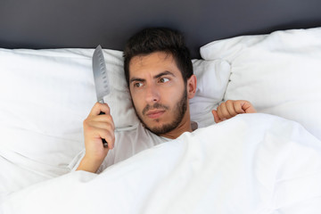 A young man is hiding in bed under a blanket at home. Fear and surprise are visible in his eyes. He...