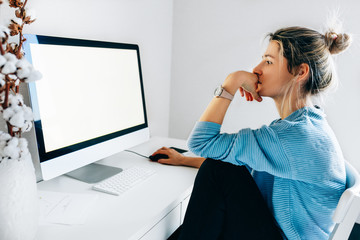 Side view image of freelancer businesswoman using a desktop computer in an office. Young blonde woman sits indoors at home working on the computer with a blank screen for your text message or content. - Powered by Adobe